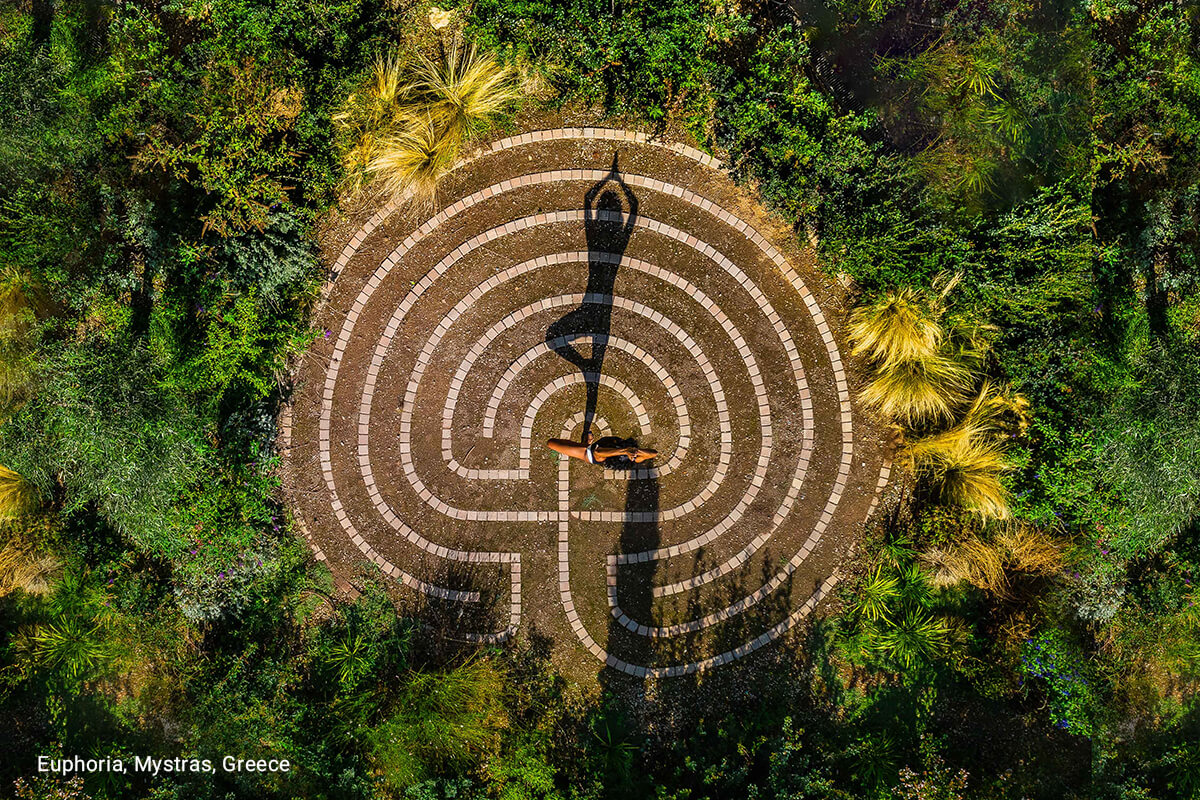 A beautiful outdoor labyrinth surrounded by lush greenery, the pattern is circular leading to the center. Sunlight is casting shadows on the ground.