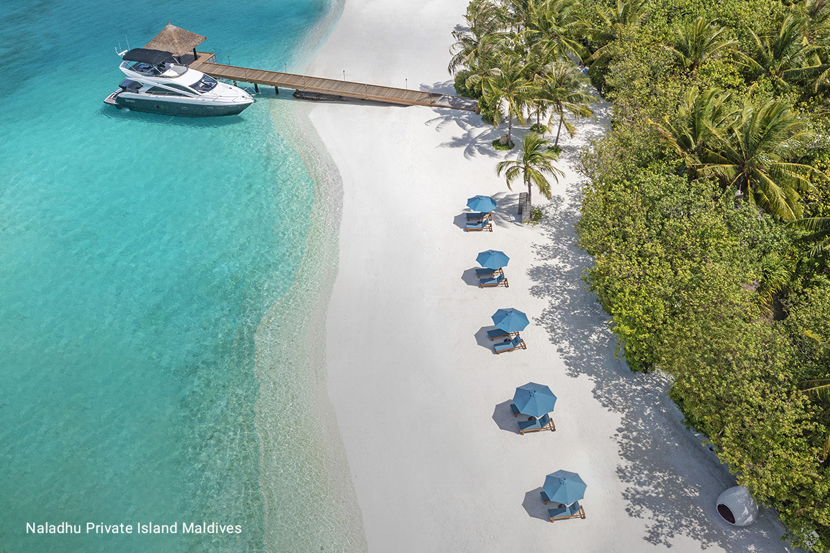 An aerial view of a beautiful tropical island beach in the Maldives. A boat is docked at the end of a boardwalk surrounded by pristine white sand and turquoise blue water.