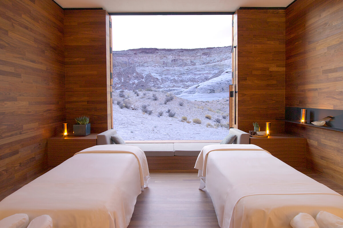 A spacious and luxurious spa treatment room at Amangiri resort in Utah. The room features a minimalist design with wooden walls and floors, and large windows that offer views of the surrounding desert landscape. In the center of the room, there are two plush massage tables with soft white sheets and towels. By the windos, there is a built-in bench with plush cushions for seating. 