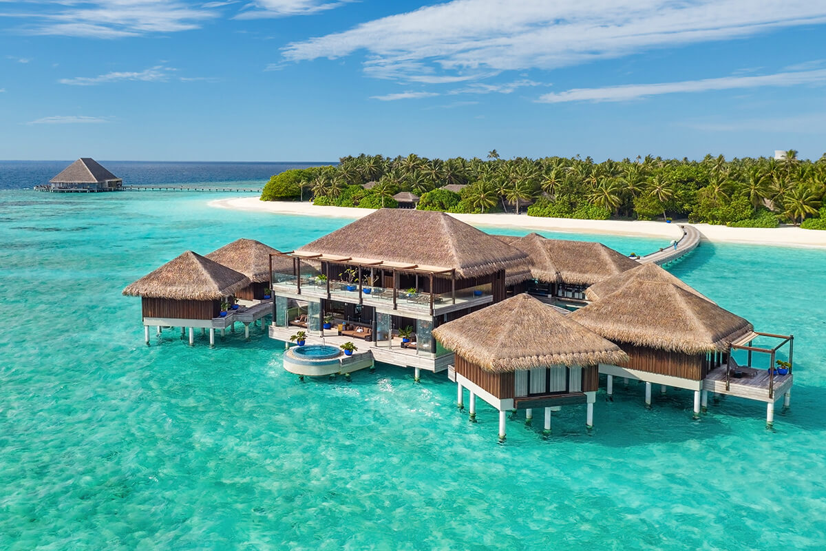 A stunning tropical island resort in the Maldives with pristine white sand beaches and crystal clear turquoise waters. The resort features overwater bungalows and villas with private pools, surrounded by lush greenery and palm trees. 