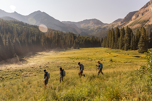 A group of hikers walking on a dirt path with a mountain range in the background. The hikers are all dressed in athletic wear and carrying backpacks, with some using trekking poles. 